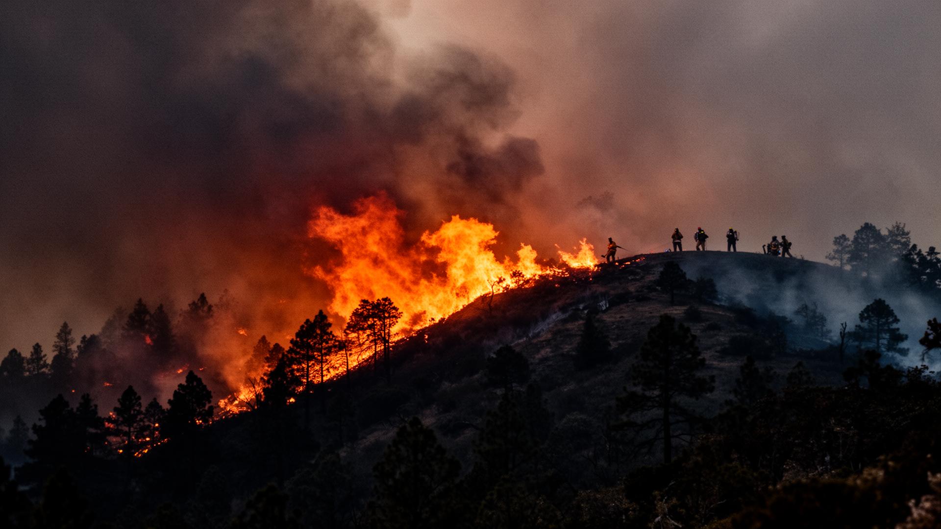 A wildfire burns along a forested ridge at dusk as firefighters watch from the crest.